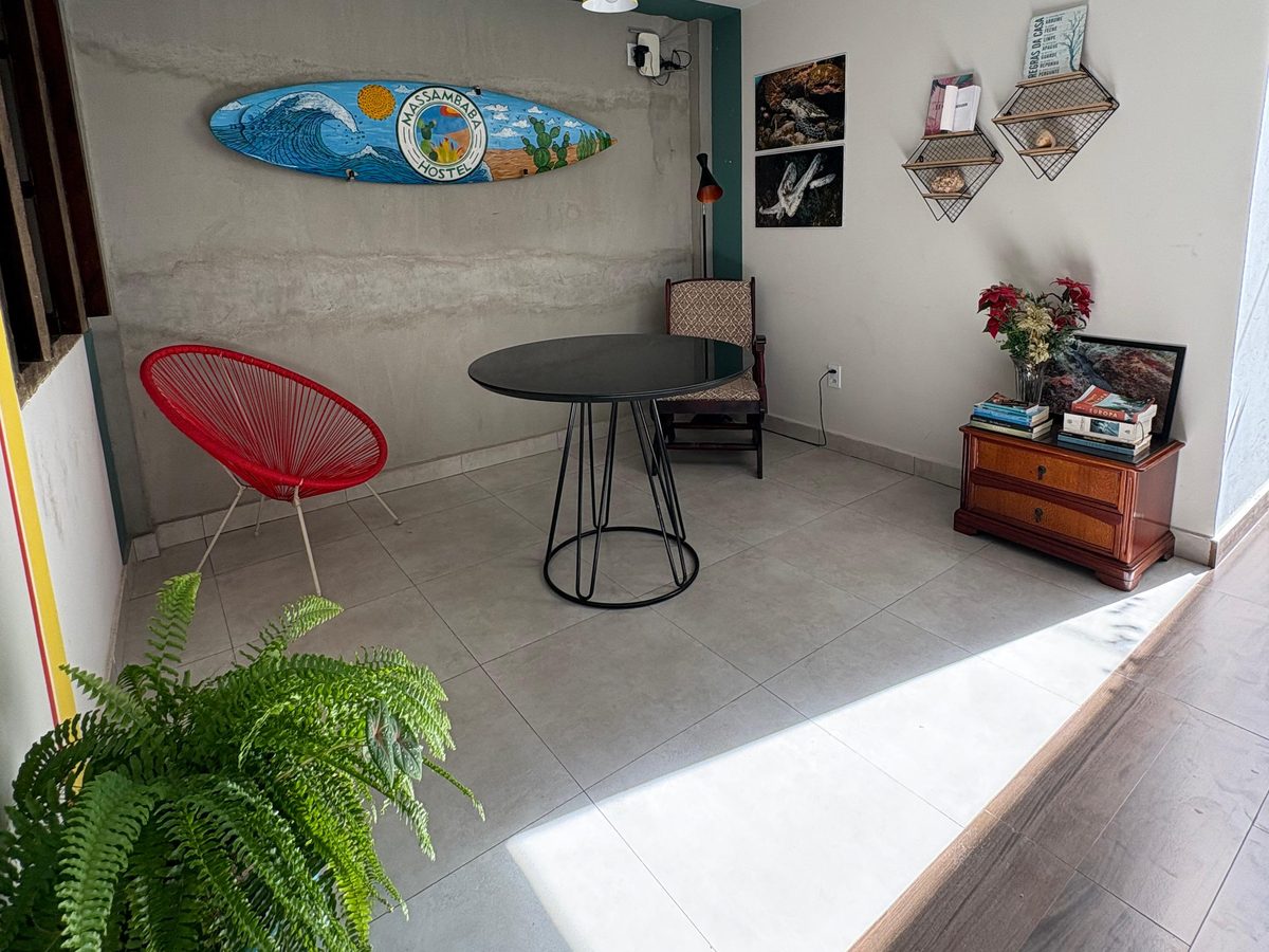 Sala de home office do Massambaba Hostel vista da porta, com mesa redonda, cadeira vermelha, cômoda com livros e a luz do sol entrando pelo piso