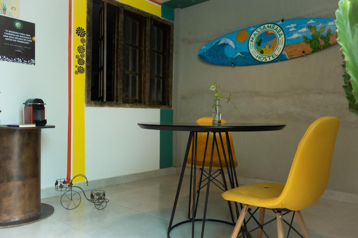 Sala de home office do Massambaba Hostel: mesa redonda preta, cadeiras amarelas e prancha pintada com o logo do hostel na parede