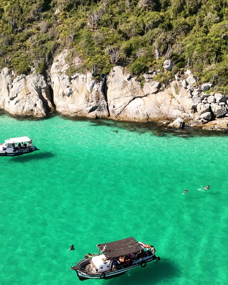 Vista aérea do barco ancorado em água verde e transparente na Enseada do Guriri, sob o costão de pedra