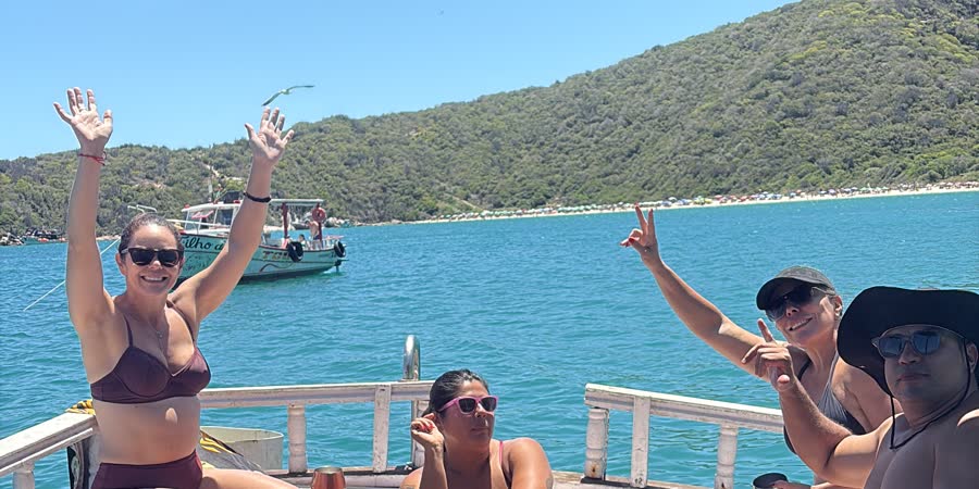Grupo de pessoas curtindo o dia no convés do barco, com o mar turquesa e o morro verde ao fundo